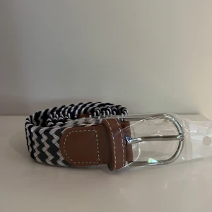 Vitt/Mörkblått flätat bälte  - Snyggt flätat bälte i blått, vitt och svart med bruna läderdetaljer och silverspänne. Perfekt accessoar för att lyfta din outfit med en sportig och stilren touch. Passar både jeans och chinos.