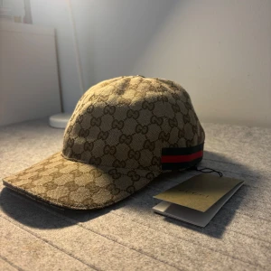 Beige keps från Gucci - Skicka dm för prisförslag/video! Snygg beige keps från Gucci med klassiskt GG-monogram över hela kepsen och en ikonisk röd-grön rand på sidan. Kepsen har böjd skärm och justerbar passform. Perfekt accessoar för en trendig look. Storlek ”M”