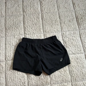 Svarta träningsshorts från Asics - Svarta träningsshorts från Asics med elastisk midja och snörning. Lätta och luftiga, perfekta för löpning eller gym. Klassisk design utan mönster och med Asics-logga på ena benet.