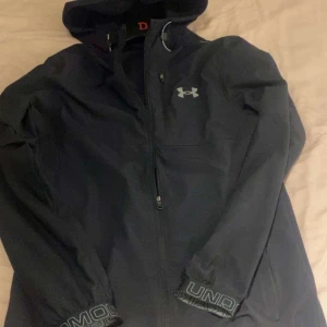 Svart vindjacka från Under Armour - Svart vindjacka från Under Armour med huva och dragkedja framtill. Jackan har logga på bröstet och tryckt text på ärmsluten. Perfekt för blåsiga dagar och sportiga outfits. Skriv och fråga på via intresse vi kommer säkert komma fram till något