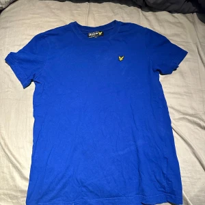 Blå t-shirt från Lyle & Scott - Snygg blå t-shirt från Lyle & Scott med klassisk rund hals och gul logga broderad på bröstet. Enkel och stilren design, perfekt till jeans eller shorts. Tillverkad i mjuk bomull för skön känsla hela dagen.