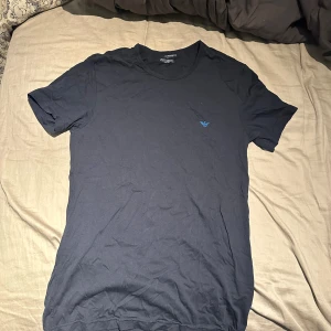 Mörkblå t-shirt från Emporio Armani - Snygg mörkblå t-shirt från Emporio Armani med diskret logga på bröstet. Tillverkad i 100% bomull för en skön och mjuk känsla. Klassisk passform och rund halsringning, perfekt till jeans eller shorts.