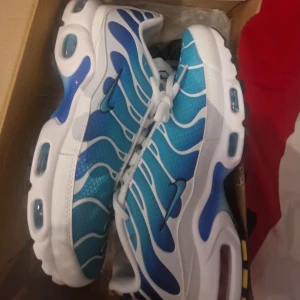 Nike Air Max Plus blå och vita sneakers - Säljer ett par Nike Air Max Plus sneakers i blått och vitt med vågigt mönster och vita snören. Skorna har synlig Air Max-dämpning i sulan och detaljer i svart och gult undertill. Perfekta för dig som gillar sportig stil.