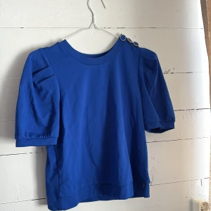 Blå topp med puffärm och knappar på axeln - Säljer en blå topp med korta puffärmar och  guldknappar på ena axeln. Väldigt mjukt och skönt material! Knappt använd så i nyskick! 