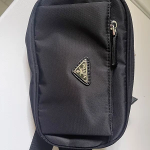 Svart axelväska från Prada - Säljer en stilren svart axelväska från Prada med klassisk trekantig logotyp framtill. Väskan har dragkedja upptill och ett extra fack med dragkedja på framsidan. Perfekt storlek för det viktigaste och enkel att bära med justerbar rem.