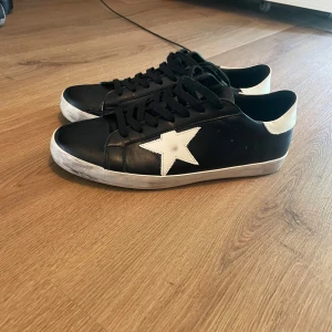 Golden Goose Super-Star svarta sneakers - Svarta Golden Goose Super-Star sneakers i skinn med vit stjärndetalj på sidan och vit hälkappa med logga. Klassisk rund tå, svarta skosnören och vit sula med lätt sliten look. Perfekta för dig som gillar streetstyle och vill sticka ut med en ikonisk sneaker. Vet ej om skorna är äkta eller inte men är fortfarande i riktigt bra kvalité. 