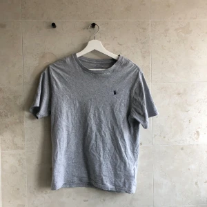 Grå t-shirt från Polo Ralph Lauren - Klassisk grå t-shirt från Polo Ralph Lauren med rund hals och korta ärmar. T-shirten har en liten broderad logga på bröstet och är gjord i mjuk bomull. Perfekt basic-plagg med clean look och skön passform.