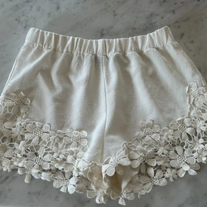 Vita shorts med spetsdetaljer - Superfina vita shorts med bred resår i midjan och dekorativa spetsblommor längs bensluten. Mjukt tyg som känns bekvämt och somrigt. Perfekta för varma dagar när du vill ha något både gulligt och stilrent.
