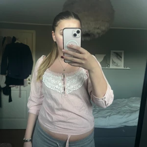 Ljusrosa blus med spetsdetaljer - Säljer en ljusrosa blus med tunna vita ränder och vackra spetsdetaljer över bysten. Blusen har trekvartsärm, knappar framtill och en feminin, romantisk känsla. Perfekt för dig som gillar söta detaljer och mjuka färger.