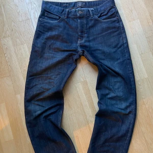 Mörkblå jeansbyxor - Säljer ett par klassiska mörkblå jeansbyxor med fem fickor och raka ben. Byxorna har normal passform och stängs med knapp och dragkedja. Perfekta till vardags och enkla att matcha med olika stilar.