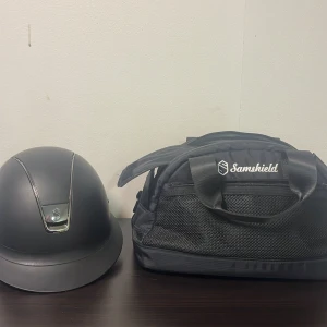 Svart ridhjälm från Samshield - Snygg svart ridhjälm från Samshield med stilrena kromdetaljer och klassisk form. Hjälmen har en matt finish och ventilerande inslag för extra komfort. Perfekt för dig som vill ha både säkerhet och stil i stallet. Man får även med hjälm väska