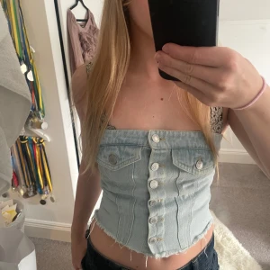 Populär Jeans tube top/korsett - Säljer en croppad ljusblå korsettopp i jeanstyg med snygga knappar framtill och rå kant nertill. Har knappt använt toppen💞 vet ej vilket märke den kommer ifrån 
