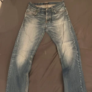 Blå jeans från Nudie Jeans Co - Säljer ett par klassiska blå jeans från Nudie Jeans Co med snygga slitningar och orangea sömmar. Jeansen har fem fickor och raka ben, perfekt för en avslappnad stil. Midjan är normal och passformen är straight. Finns en defekt som är med på sista bilden. Nyansen kan skilja sig lite åt. 