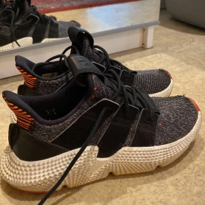 Adidas Prophere sneakers med svart och orange detaljer - Säljer ett par Adidas Prophere sneakers i svart med stickad ovandel och orange detaljer på hälen. Skorna har en kraftig vit sula, svarta snören och diskreta röda prickar i tyget. Perfekta för dig som vill sticka ut med en unik sneaker. Använda några få gånger 