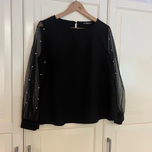Svart blus med pärldetaljer - En svart blus med transparenta långärmade ärmar i mesh och pärldetaljer längs ärmarna. Passar även Stl.M.