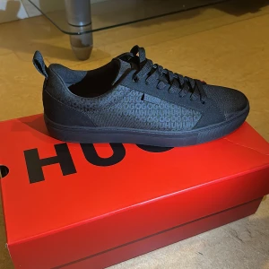 Svarta sneakers från Hugo Boss - Säljer ett par helsvarta sneakers från Hugo Boss med diskret logga på sidan. Skorna har snörning, rund tå och platt sula. Perfekta för en clean och stilren look.