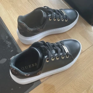 Svarta sneakers från Guess med logga - Snygga svarta sneakers från Guess med vita platta sulor och guldfärgade detaljer. Skorna har snörning och Guess-logga framtill samt mönstrad kant med logotyp runt hälen. Perfekta för dig som gillar stilrena men ändå trendiga skor.
