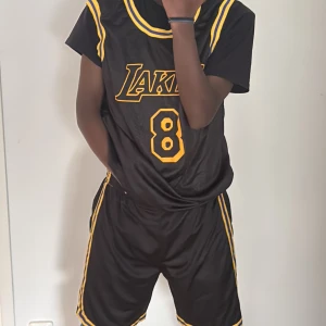 Lakers basket tröja och shorts svart/gul - Snygga basketshorts från Lakers i svart med gula och lila ränder längs sidorna. Klassisk loose fit och elastisk midja för skön passform. Perfekta för basket eller streetwear. Matchar med Bryant-tröja för en komplett look.