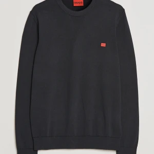 Svart stickad tröja från Hugo - Snygg svart stickad tröja från Hugo med rund halsringning och diskret röd logotyp på bröstet. Tröjan har långa ärmar och ribbade muddar vid ärmslut och nederkant. Perfekt för en stilren och enkel look.