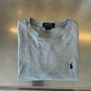 Grå t-shirt från Polo Ralph Lauren - Klassisk grå t-shirt från Polo Ralph Lauren med rund halsringning och den ikoniska broderade loggan på bröstet. Perfekt basplagg med enkel och stilren design. Den är lite liten i storleken! Skulle säga att den passar storleken M.           