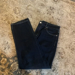 Mörkblå raka jeans - Snygga mörkblå jeans med klassisk femficksdesign och raka ben. Jeansen har silverfärgade nitar och knapp, samt kontrastsömmar. Perfekta för en stilren och enkel look.