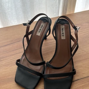 Svarta sandaler med klack från NA-KD - Säljer ett par stilrena svarta sandaler från NA-KD med smala remmar och fyrkantig tå. Skorna har en elegant klack och justerbar rem runt ankeln. Perfekta för att lyfta vilken outfit som helst.