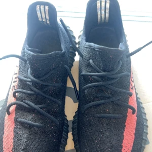 Adidas Yeezy Boost 350 V2 Black/Red - Säljer ett par Adidas Yeezy Boost 350 V2 i svart med röd rand och texten SPLY-350 på sidan. Skorna har en stickad ovandel, platt sula och klassisk Yeezy-design. Perfekta för dig som vill sticka ut med en ikonisk sneaker.
