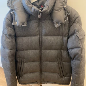 Grå Moncler pufferjacka med huva - Säljer en grå pufferjacka från Moncler med avtagbar huva och dragkedja framtill. Jackan har två fickor med dragkedja, en extra ficka på ärmen med Moncler-logga och quiltad design. Perfekt för kalla dagar och riktigt snygg till streetwear.