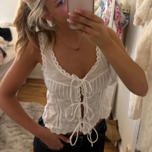 Vit broderad blus med snörning - Superfin vit blus med broderade detaljer och spetskant. Toppen har v-ringning, breda axelband och snörning framtill som ger en romantisk vibe. Perfekt till jeans eller kjol för en söt look. Materialet känns lätt och luftigt.