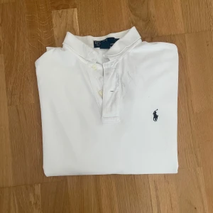 Vit pikétröja från Polo Ralph Lauren - Klassisk vit pikétröja från Polo Ralph Lauren med broderad marinblå logga på bröstet. Tröjan har korta ärmar, krage och knäppning med tre knappar. Tillverkad i 100% bomull för en skön och luftig känsla. OBS, lite smutsig i kragen, går möjligtvis bort i tvätten. 