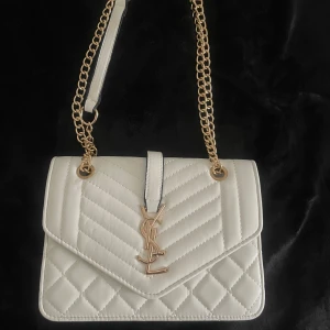 Vit quiltad axelväska från YSL - Säljer en snygg vit axelväska från YSL med quiltad design och guldiga kedjor. Väskan har YSL-logga framtill, magnetlås och ett fack med dragkedja inuti. Aldrig använd och i perfekt skick, får plats med väldigt mycket. Pris kan diskuteras 