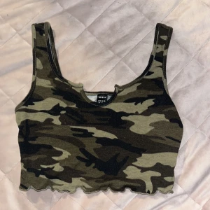 Camouflage croppad linnetopp Shein XS - Säljer en croppad linnetopp från Shein i storlek XS med camouflage-mönster i gröna och bruna toner. Toppen har ärmlös design, liten v-ringning och vågig kant nertill. Perfekt för dig som gillar streetstyle och vill sticka ut.