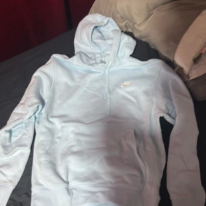 Ljusblå hoodie från Nike - Säljer en ljusblå hoodie från Nike med klassisk logga broderad på bröstet. Tröjan har huva och en stor magficka framtill. Perfekt för en avslappnad stil och enkel att matcha med jeans eller mjukisbyxor.