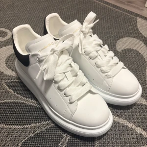 Alexander McQueen - Säljer ett par vita sneakers från Alexander McQueen med svart hälparti och kraftig plattformssula. Skorna har snörning och är tillverkade i skinn med detaljer i mocka. Klassisk och stilren design med logga i guld på hälen.