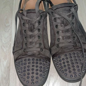 Christian Louboutin gråa sneakers med nitar - Unika svarta sneakers från Christian Louboutin i mocka med coola svarta nitar på tån och klassisk röd sula. Skorna har snörning och rund tå, perfekt för dig som vill sticka ut med en lyxig streetstyle-look.
