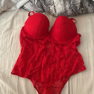 Röd spetskorsett med bygel och knäppning - Säljer en röd body i spets med bygelkupor och justerbara axelband. Modellen har knäppning i grenen och vacker blommig spets över hela plagget. Perfekt för att känna sig extra fin.