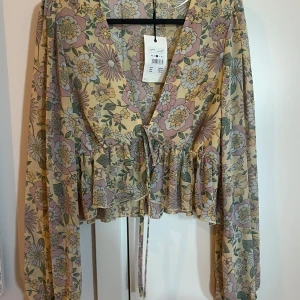 Blommig blus med volang från Bik Bok - Supersöt blommig blus från Bik Bok i pastelliga toner av beige, rosa och grön. Blusen har långa ärmar, volangdetalj nedtill och knyt i midjan. Perfekt för dig som gillar romantisk stil och mjuka färger.