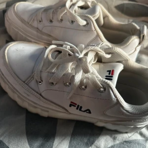 Vita sneakers från Fila - Chunky vita sneakers från Fila med klassisk logga på sidan och plösen. Skorna har snörning, rund tå och tjock sula. Perfekta för dig som gillar en sportig och trendig stil. Tillverkade i syntetmaterial med textilfoder.