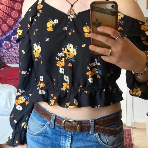 Blommig croppad offshoulder topp från H&M, M - Superfin svart croppad blus med gula och vita blommor. Offshoulder-modell med långa ärmar, volangdetaljer och knyt framtill. Axelbanden är justerbara. Tunnt svalt material. Perfekt för sommaren!