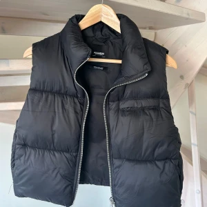 Svart dunväst från Pull&Bear - Säljer en svart dunväst från Pull&Bear med hög krage och dragkedja framtill. Västen har en quiltad design och en bröstficka med dragkedja. Perfekt att slänga över en hoodie eller tröja för extra värme.