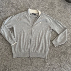 Grå ziptröja från Polo Ralph Lauren - Snygg grå ziptröja från Polo Ralph Lauren med vit ribbad krage och ärmslut. Tröjan har dragkedja framtill, långa ärmar och diskret broderad logga på bröstet. Perfekt för en stilren och avslappnad look.