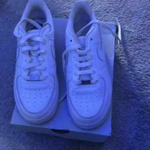 Nike Air Force 1 vita sneakers - Klassiska vita Nike Air Force 1 sneakers med låg profil och rund tå. Skorna har snörning och är tillverkade i skinn med perforerade detaljer på tån för extra ventilation. Perfekta för en clean och stilren look.