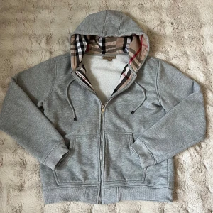 Grå hoodie med dragkedja från Burberry - Säljer en grå hoodie från Burberry med klassiskt rutigt foder i huvan. Tröjan har dragkedja framtill, två fickor och justerbar huva med snören. Perfekt för en stilren och avslappnad look. Storlek L