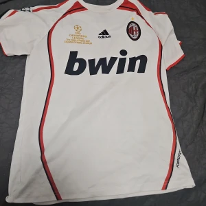 AC Milan bortatröja Maldini 3 Champions League - Vit AC Milan bortatröja med röda och svarta detaljer, Adidas-logga och Bwin sponsortryck. Champions League-märken på ärmarna och Maldini 3 på ryggen. Klassisk fotbollströja med klubbmärke på bröstet.