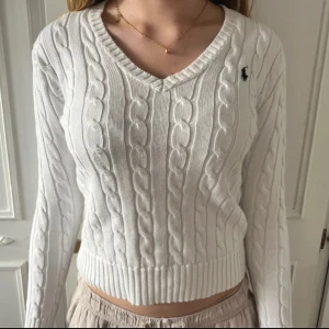 Vit kabelstickad tröja från Polo Ralph Lauren - Säljer en klassisk vit kabelstickad tröja från Polo Ralph Lauren med v-ringning och broderad logga på bröstet. Tröjan är croppad och har långa ärmar samt ribbade muddar. Perfekt för en stilren och tidlös look.
