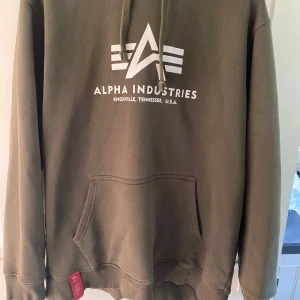 Olivgrön hoodie från Alpha Industries - Säljer en olivgrön hoodie från Alpha Industries med vit logga och text på bröstet. Tröjan har huva med dragsko och en stor magficka framtill. Perfekt för dig som gillar streetwear och vill ha en bekväm och snygg tröja.
