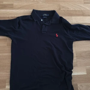Svart pikétröja Polo Ralph Lauren S - Klassisk svart pikétröja från Polo Ralph Lauren i bomull, med röd broderad logga på bröstet. Tröjan har krage, knappar vid halsen och en custom fit-passform. Perfekt för en stilren och sportig look.