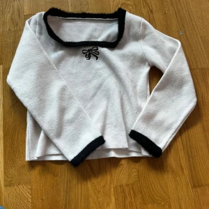 Vit långärmad topp med svarta detaljer och rosett - Säljer en vit långärmad topp med svarta kanter vid halsringning och ärmslut. Söt liten svart rosett framtill. Fyrkantig ringning och mjukt material som känns skönt mot huden. Perfekt för dig som gillar stilrena men ändå unika plagg.