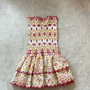 Vit och röd klänning från Design by Amor - Supersöt axelbandslös klänning från Design by Amor med volangkant och smock upptill. Klänningen har ett färgglatt mönster i rött och gult på vit botten samt volanger nertill. Perfekt för sommarens alla tillfällen!