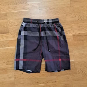 Rutig badshorts med snörning - Snygga badshorts med svart, grått och rött rutmönster. De har elastisk midja med svart snörning och är perfekta för sommardagar vid stranden eller poolen. Klassisk passform och lätt material.